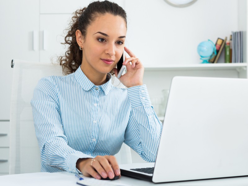 Junge Frau sitzt im Büro vor Laptop und telefoniert mit Handy