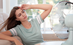 Wie kann ich meine Wohnung im Sommer ohne Luftzug kühlen? Frau sitzt mit wehenden langen Haaren auf Sofa vor einem Ventilator.