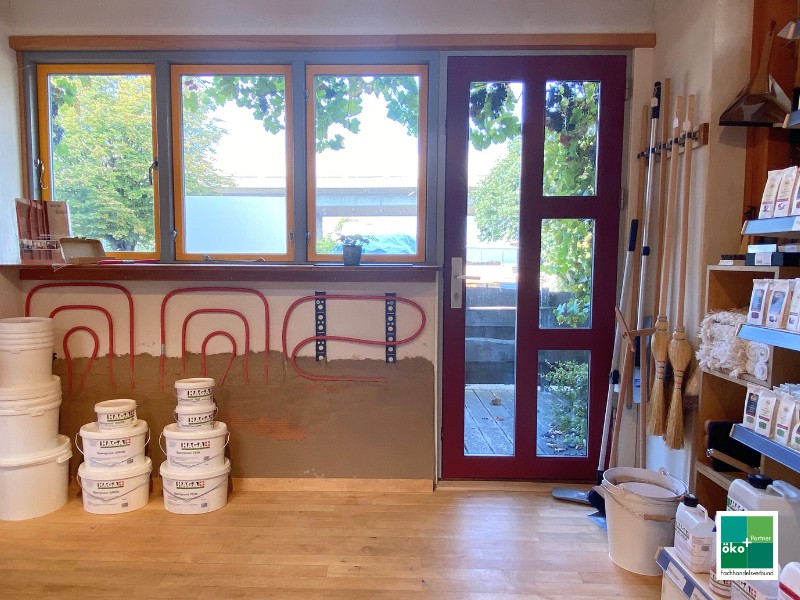 Naturbaustoffe bei ÖkoPlus-Händler im Laden: Wandheizung unter dem Fenster - teilweise mit Lehm verputzt. Eimer mit Kalkputz stehen daneben und im Regal